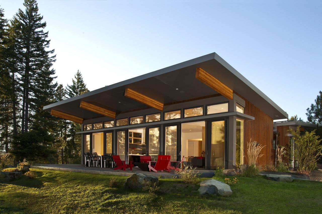  Modern cabin with an extended sloped roof, large glass windows, and a patio with red chairs, surrounded by evergreen trees.