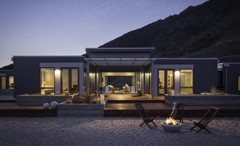 Modern house with a large open living area and wooden deck, facing a beach with a fire pit, at dusk with a dark hill in the background.