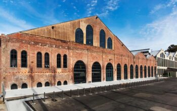 An old factory or warehouse with a long, gabled roof and numerous large arched windows, with a clean concrete plaza in front.