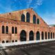 An old factory or warehouse with a long, gabled roof and numerous large arched windows, with a clean concrete plaza in front.