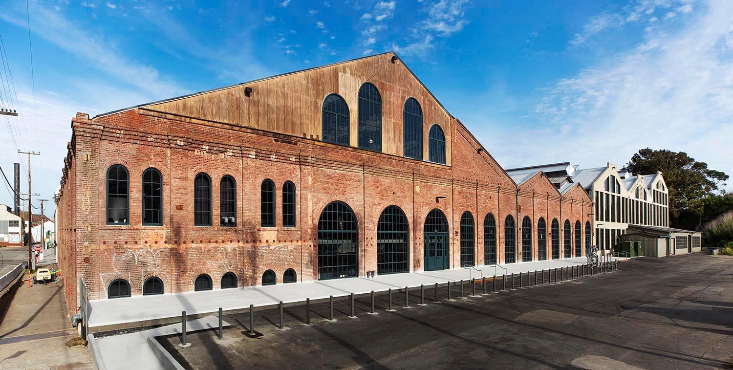 An old factory or warehouse with a long, gabled roof and numerous large arched windows, with a clean concrete plaza in front.