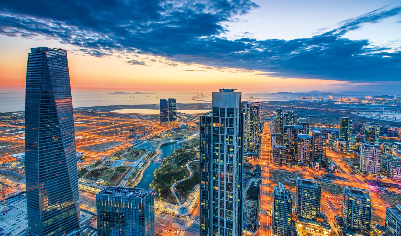  Aerial view of a modern coastal city at dusk, with illuminated skyscrapers, green spaces, and a bridge in the distance.