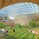 Indoor public space with a large grassy area, trees, wooden tiered seating, and a skylight roof covered in solar panels.