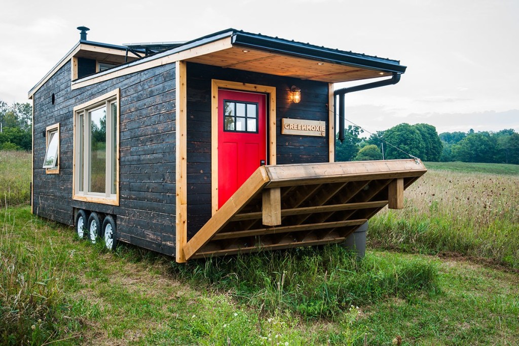 A black tiny house with a red door and wooden porch, set in a green field with trees in the background.