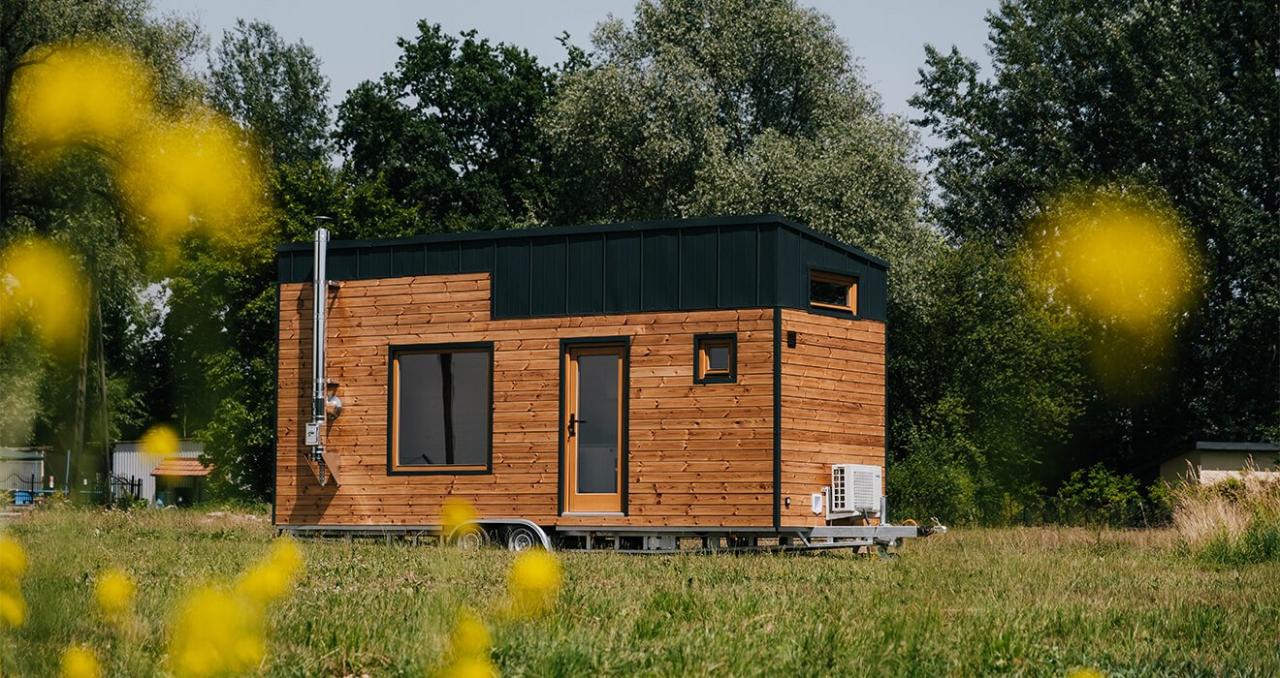 A small, modern tiny house with wood siding and a black metal roof, set in a green field under a clear sky.