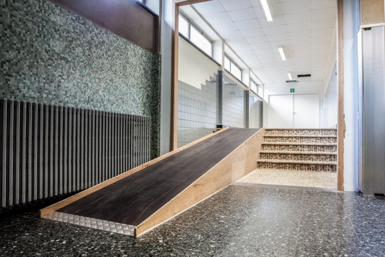 An indoor ramp with a dark surface and light wood sides, next to a set of stairs, leading towards a hallway with tiled walls and a white door.